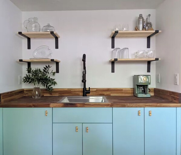 A kitchen with a wooden countertop, a sink, and cabinets. There are two shelves above the countertop holding glassware and a coffee machine.