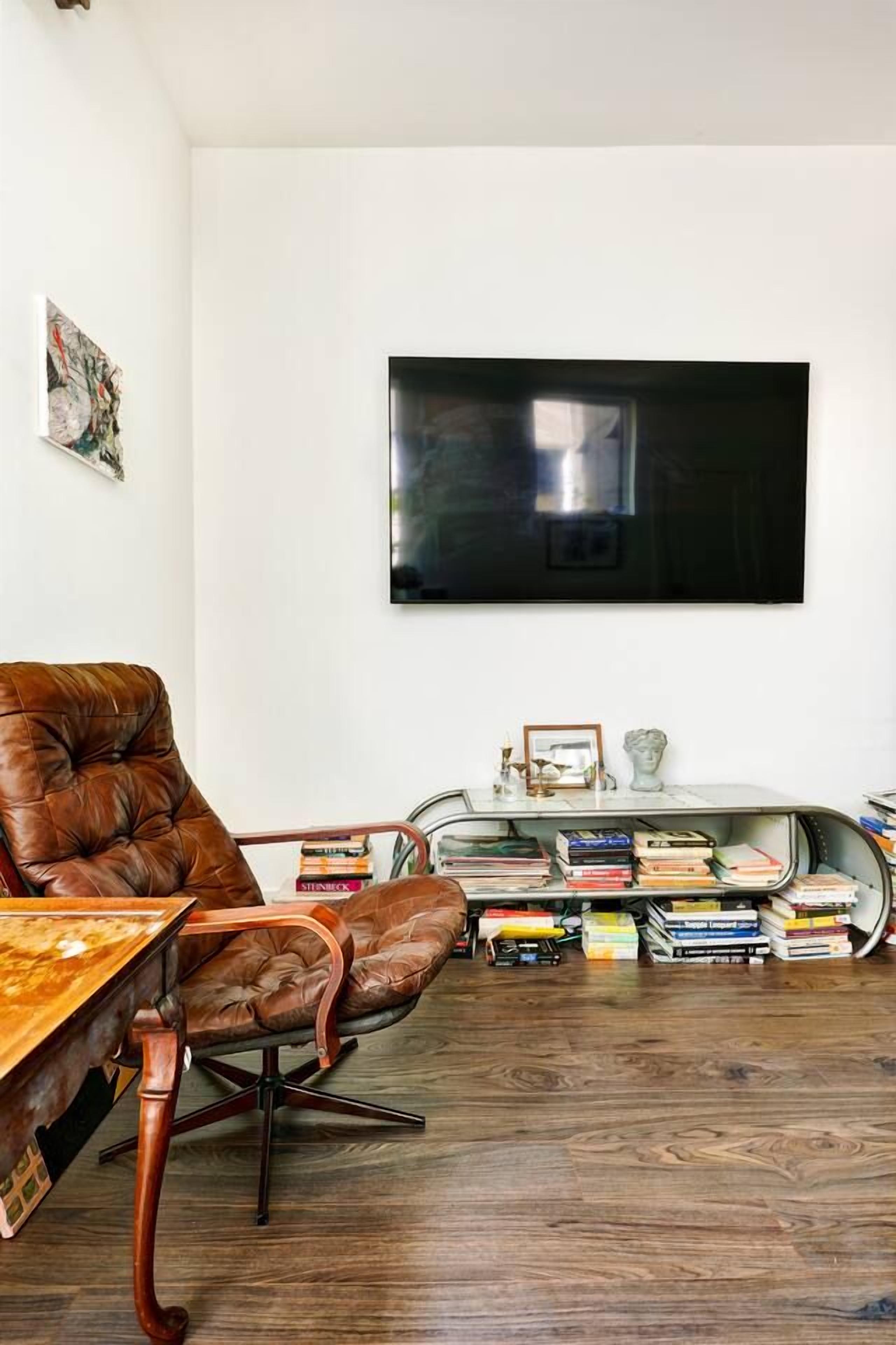 A room with a leather chair, a wooden table, a bookshelf, and a flat-screen TV mounted on the wall.