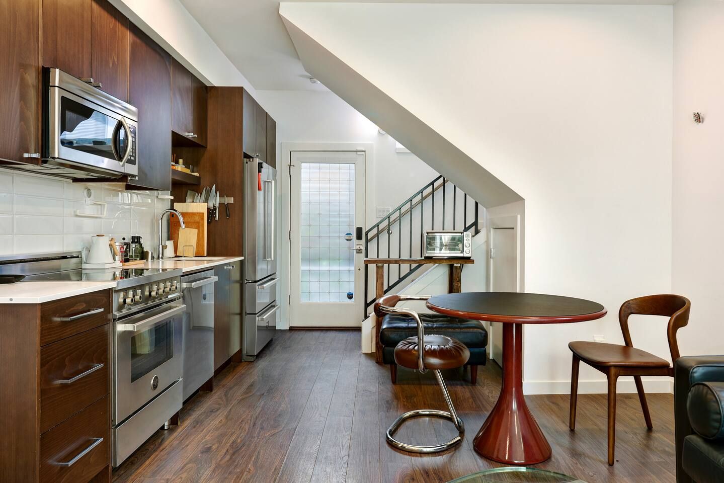 A modern kitchen with stainless steel appliances, a dining area with a round table and chairs, and a staircase leading to another level.