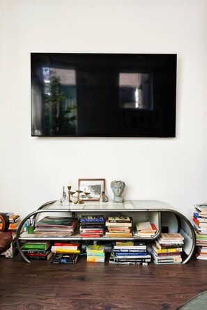 A room with a flat-screen TV mounted on the wall, a curved white console table with books and decorative items, and a wooden floor. - Artist's Townhouse in Capitol Hill w/ AC + Rooftop (Seattle)
