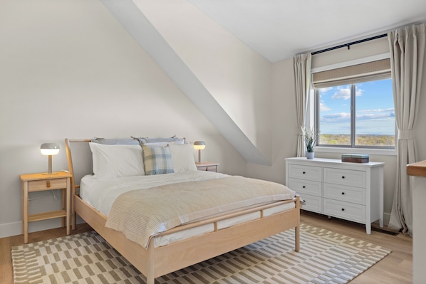 Een slaapkamer met een schuine kap, een bed met houten frame, bedlampen, een dressoir en een raam met gordijnen.