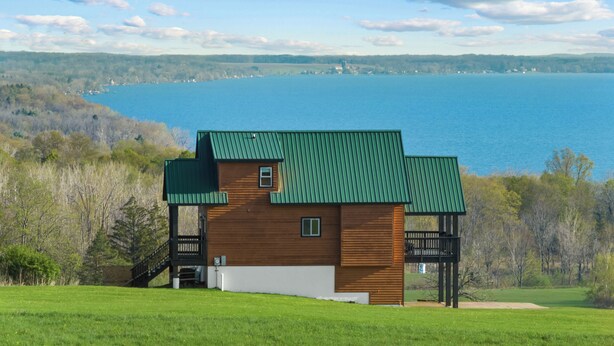 A wooden cabin with a green roof, situated on a grassy hill overlooking a large body of water. - Perched Above Cayuga ·  Sauna · Winery On-Site (Romulus)