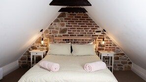 A bedroom with a sloped ceiling, exposed brick walls, a wooden headboard, two bedside tables, and a bed with striped bedding and rolled towels.
