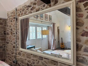 A stone-walled room with a large mirror reflecting a door, a guitar, and a lamp.