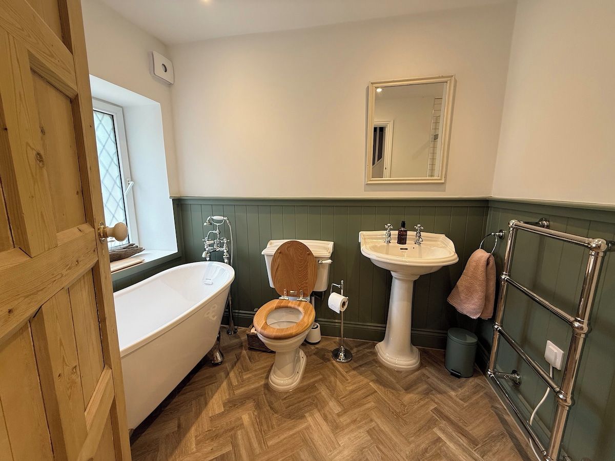 Un baño con puerta de madera, una bañera blanca, inodoro de madera, lavamanos pedestal y un perchero para toallas.