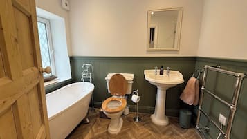 Un baño con puerta de madera, una bañera blanca, inodoro de madera, lavamanos pedestal y un perchero para toallas.