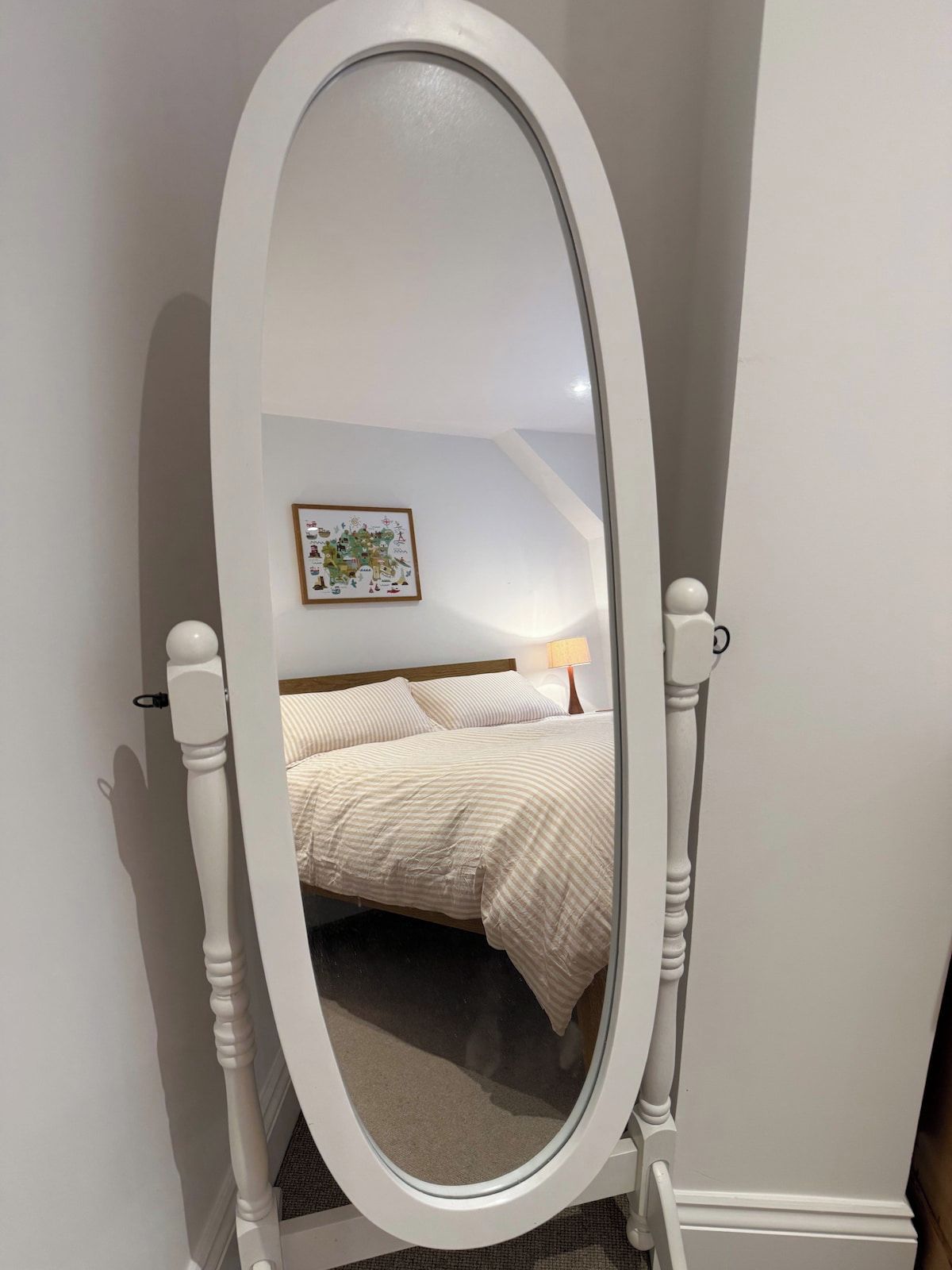 Un espejo blanco con marco ovalado en una habitación que tiene una cama, un cuadro en la pared y una mesita de noche con una lámpara.
