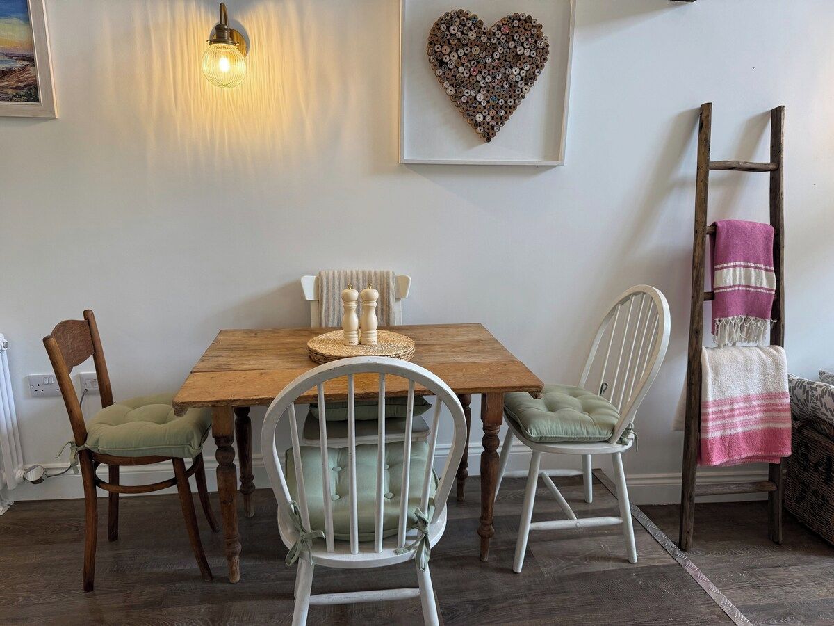 Uma área de refeições com mesa de madeira, duas cadeiras, luminária fixada na parede e uma decoração em formato de coração.
