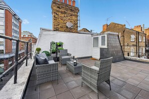 Eine Dachterrasse mit Korbmöbeln, einer weißen Wand und einem Backsteinturm im Hintergrund.