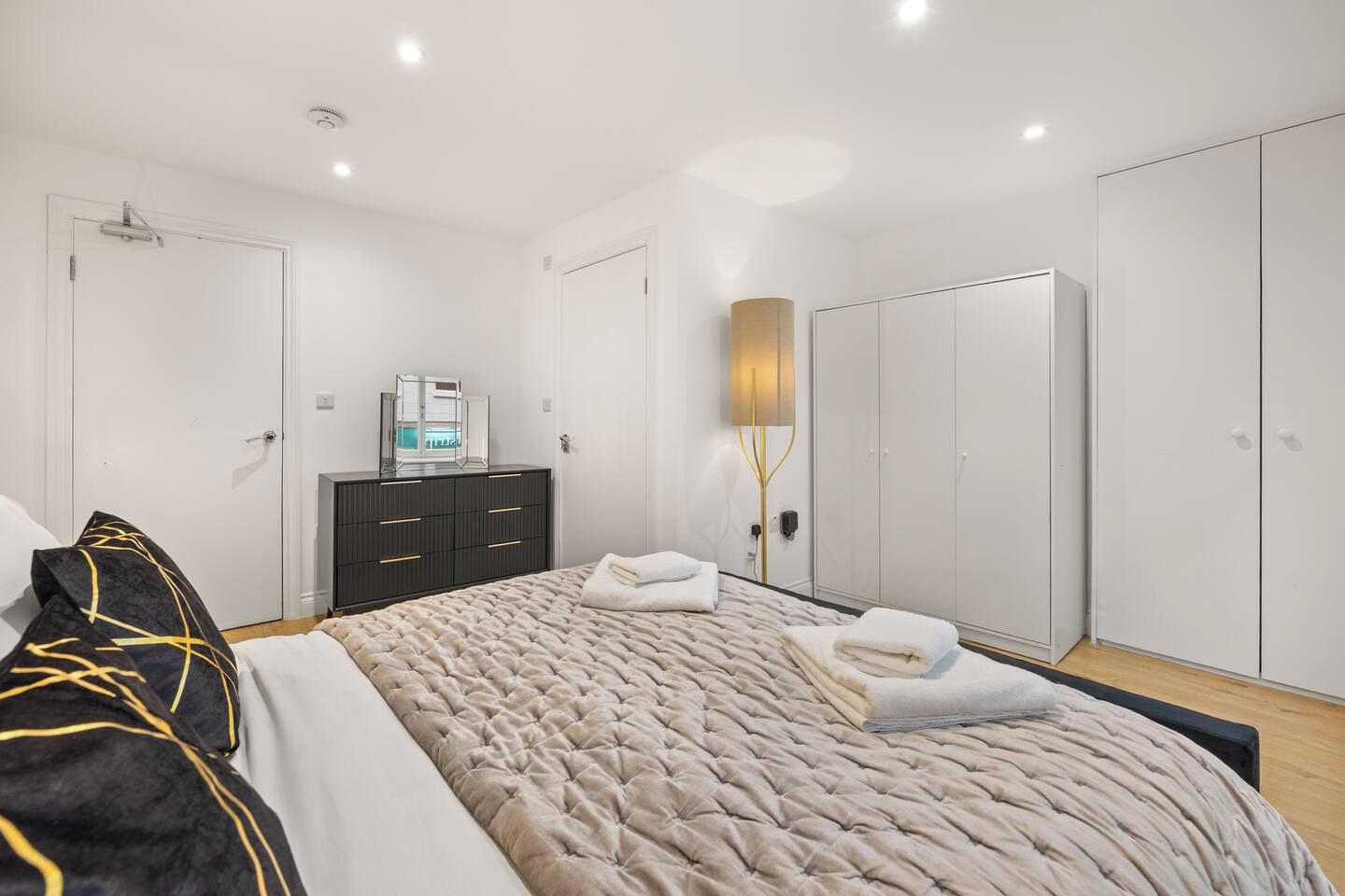 A modern bedroom with a large bed, a dresser, a wardrobe, and a bedside lamp.