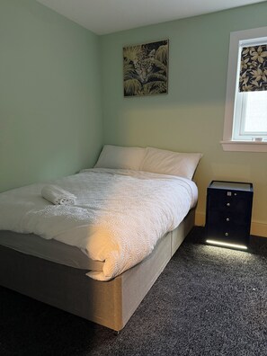 A neatly made bed with a white comforter and pillows, a small bedside table with drawers, a wall-mounted artwork, and a window with a view. - Cosy and bright spacious 3 bedroom house in a quiet cul de sac area in Culloden (Culloden)