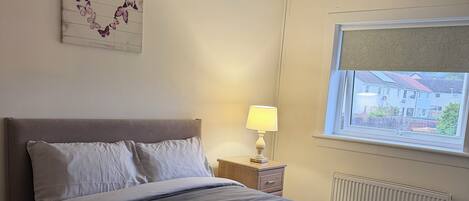 A hotel room with a bed, a nightstand with a lamp, a window with a view, and a wall decoration.