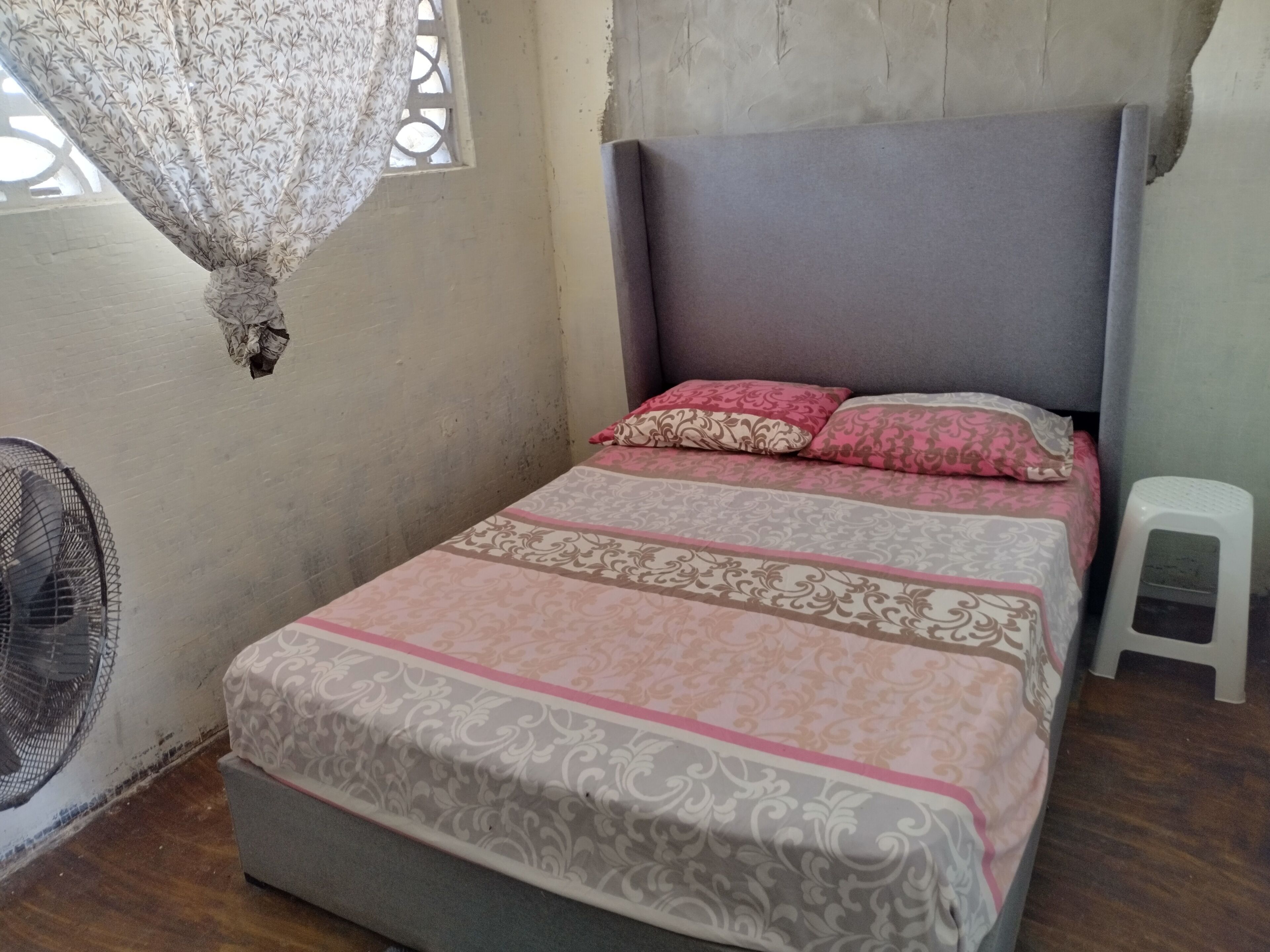 A single bed with patterned bedding, a small stool, a standing fan, and a window with a curtain.