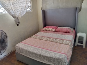 A single bed with patterned bedding, a small stool, a standing fan, and a window with a curtain. - BLUE SEA HOUSE (Playa Blanca)