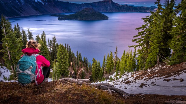 A person in a red jacket and turquoise backpack sitting on a grassy slope overlooking a lake with mountains in the background. - Cozy Queen Suite 104 –Stroll Historic Downtown, Great Restaurants & Unique Shops (Grants Pass)