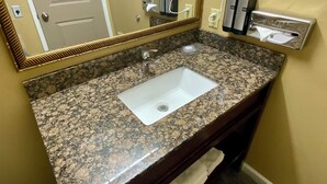 A bathroom with a granite countertop sink, a mirror, a hairdryer, and a towel rack. - Cozy Queen Suite 104 –Stroll Historic Downtown, Great Restaurants & Unique Shops (Grants Pass)