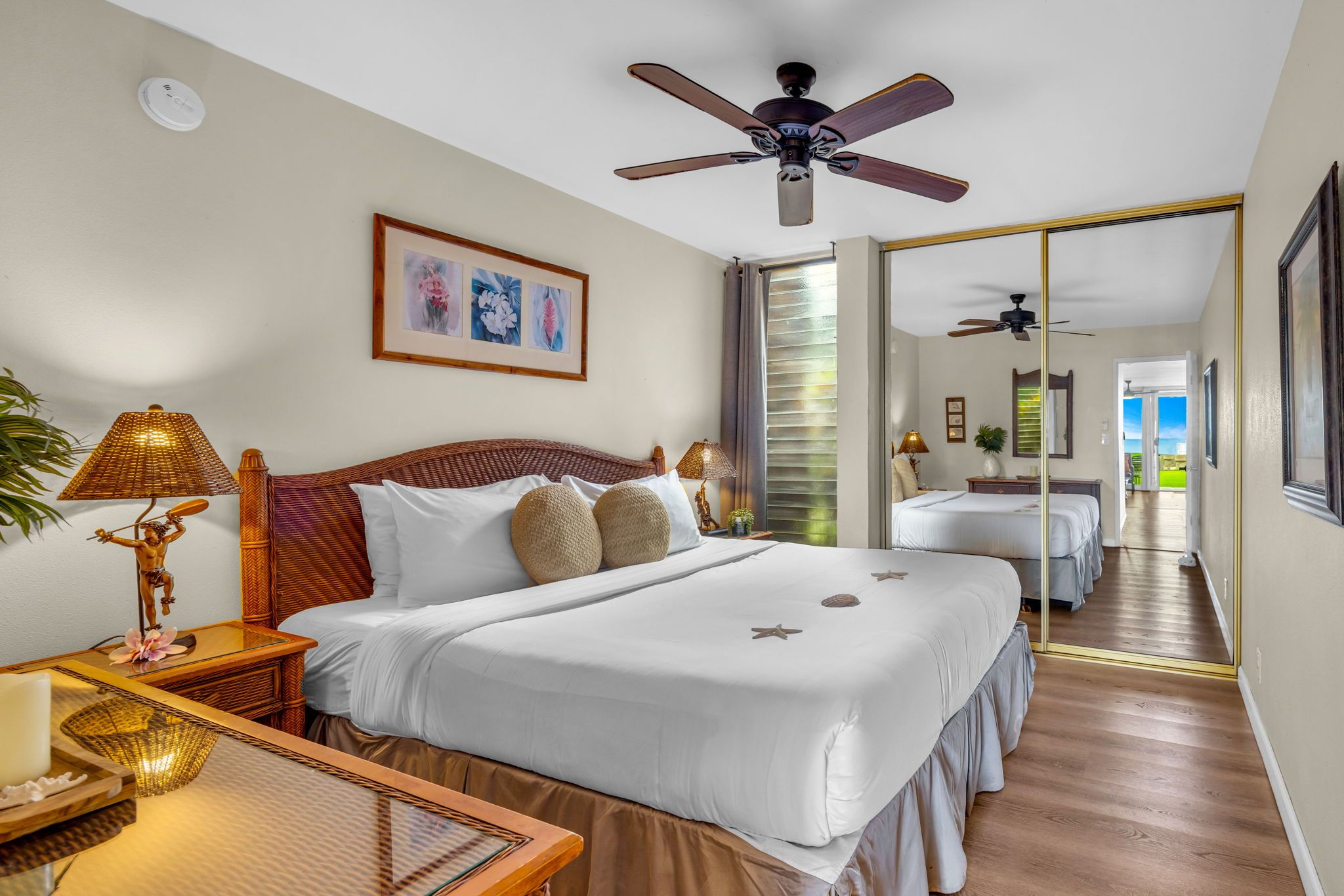 Une chambre d’hôtel avec un lit, des tables de chevet, un ventilateur de plafond, un tableau encadré et une vue sur l’extérieur, visible à travers une porte ouverte.