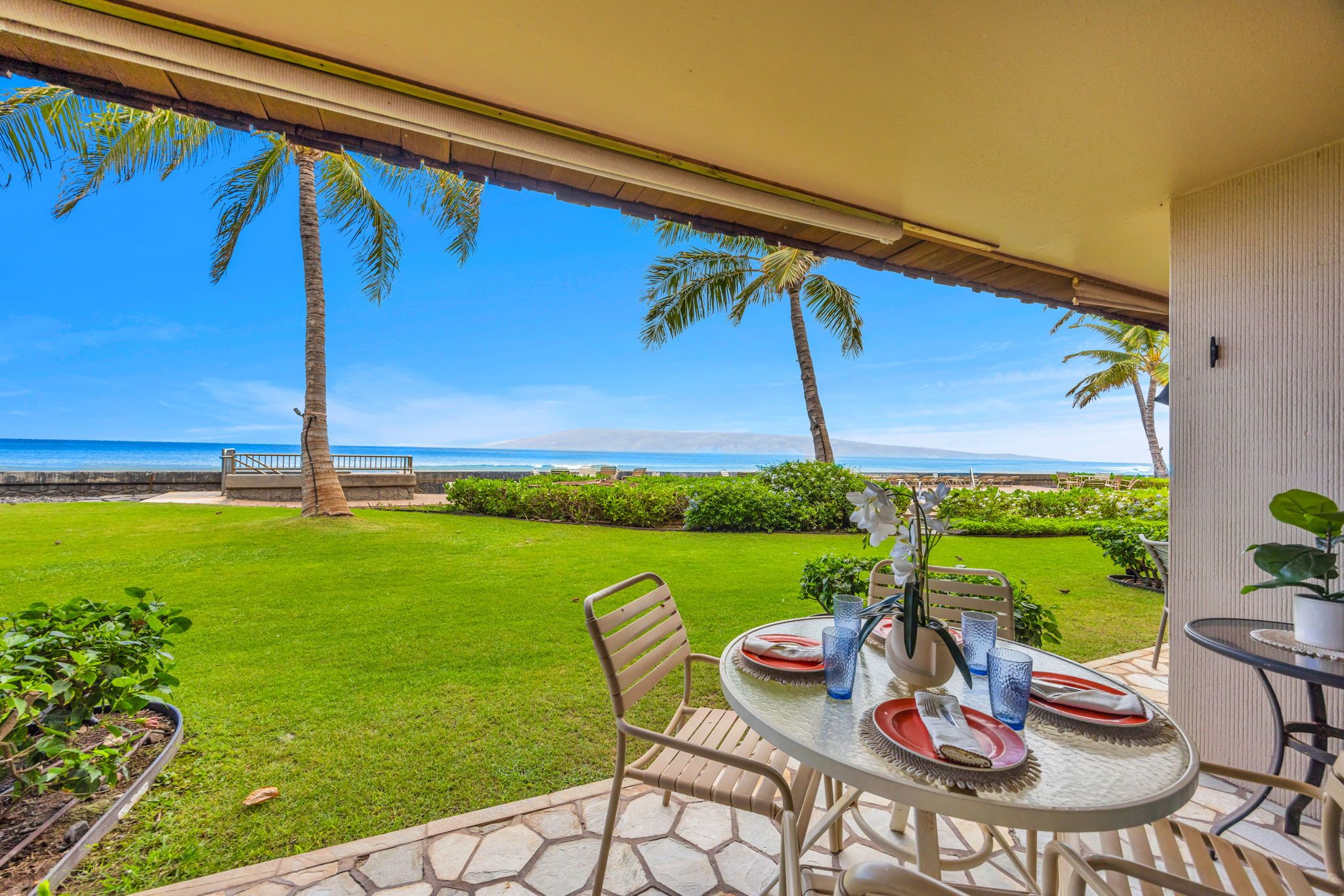 A patio with a table set for dining, overlooking a beach with palm trees and a clear sky.