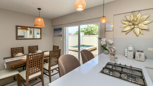 Una cucina moderna con isola centrale, zona pranzo con sedie in legno, illuminazione a sospensione e vista su una piscina esterna.