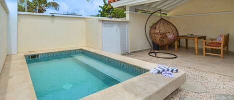 Una piscina con borde de azulejos, una zona de estar cubierta con mesa y sillas, y una silla colgante.