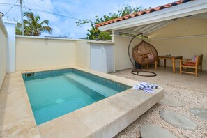 Una piscina con borde de azulejos, una zona de estar cubierta con mesa y sillas, y una silla colgante.