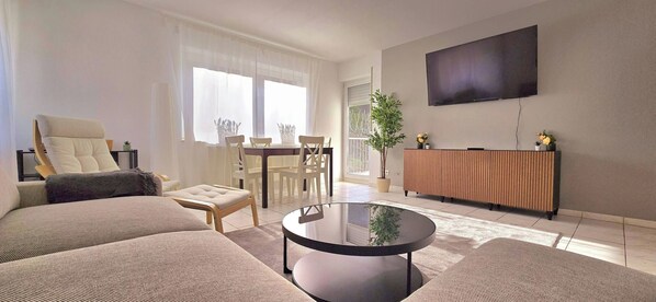 A modern living room with a sofa, armchair, dining table, chairs, a coffee table, a TV, and a potted plant. - Mosel Apartment III - MIS (Traben-Trarbach)