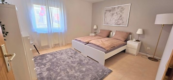 Un dormitorio con cama, mesitas de noche, lámpara, reloj y ventana con cortinas.
