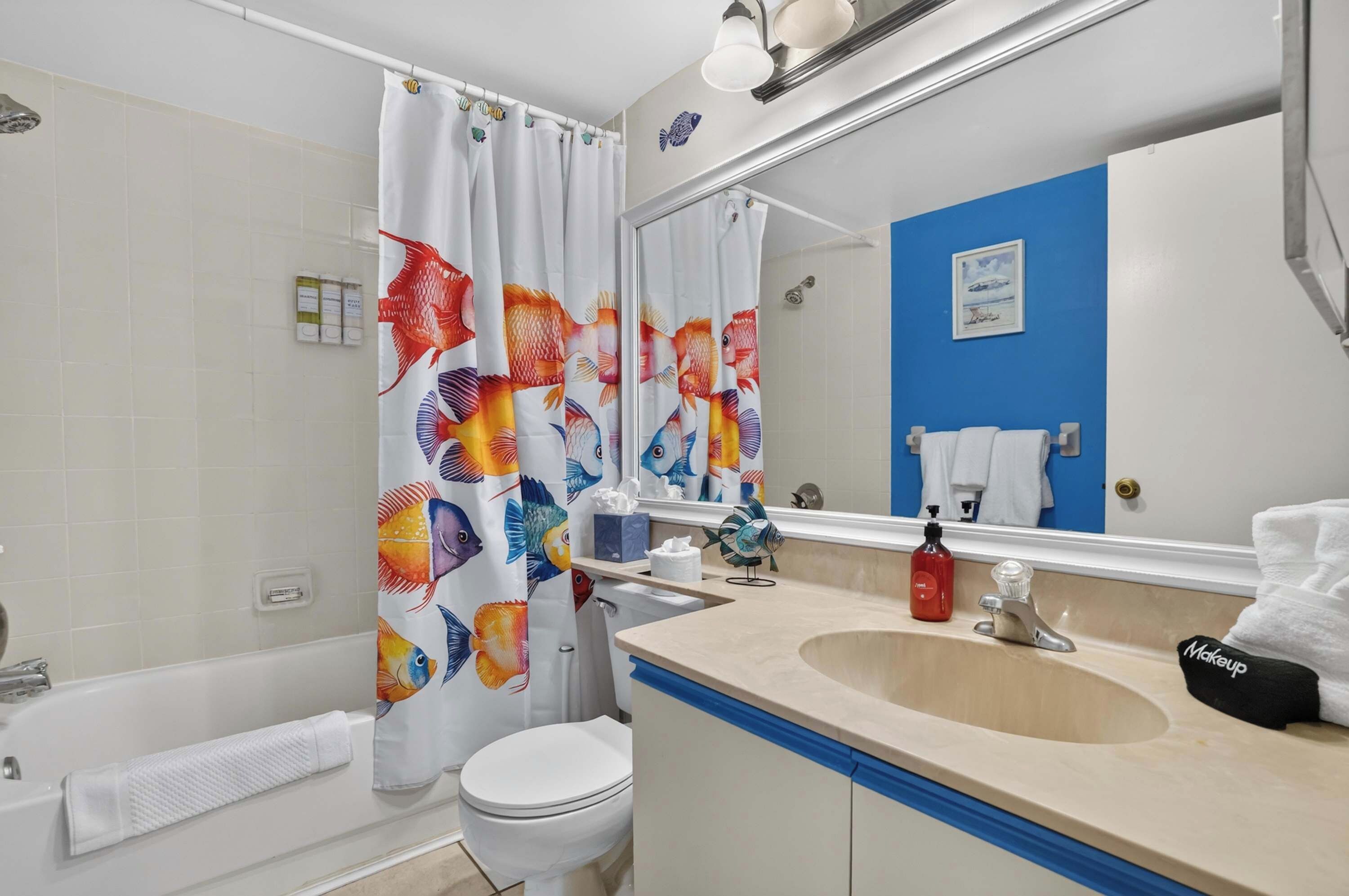Ein Badezimmer mit Badewanne, Toilette, Waschbecken und einem Spiegel, der eine blaue Wand und ein Strandbild reflektiert.
