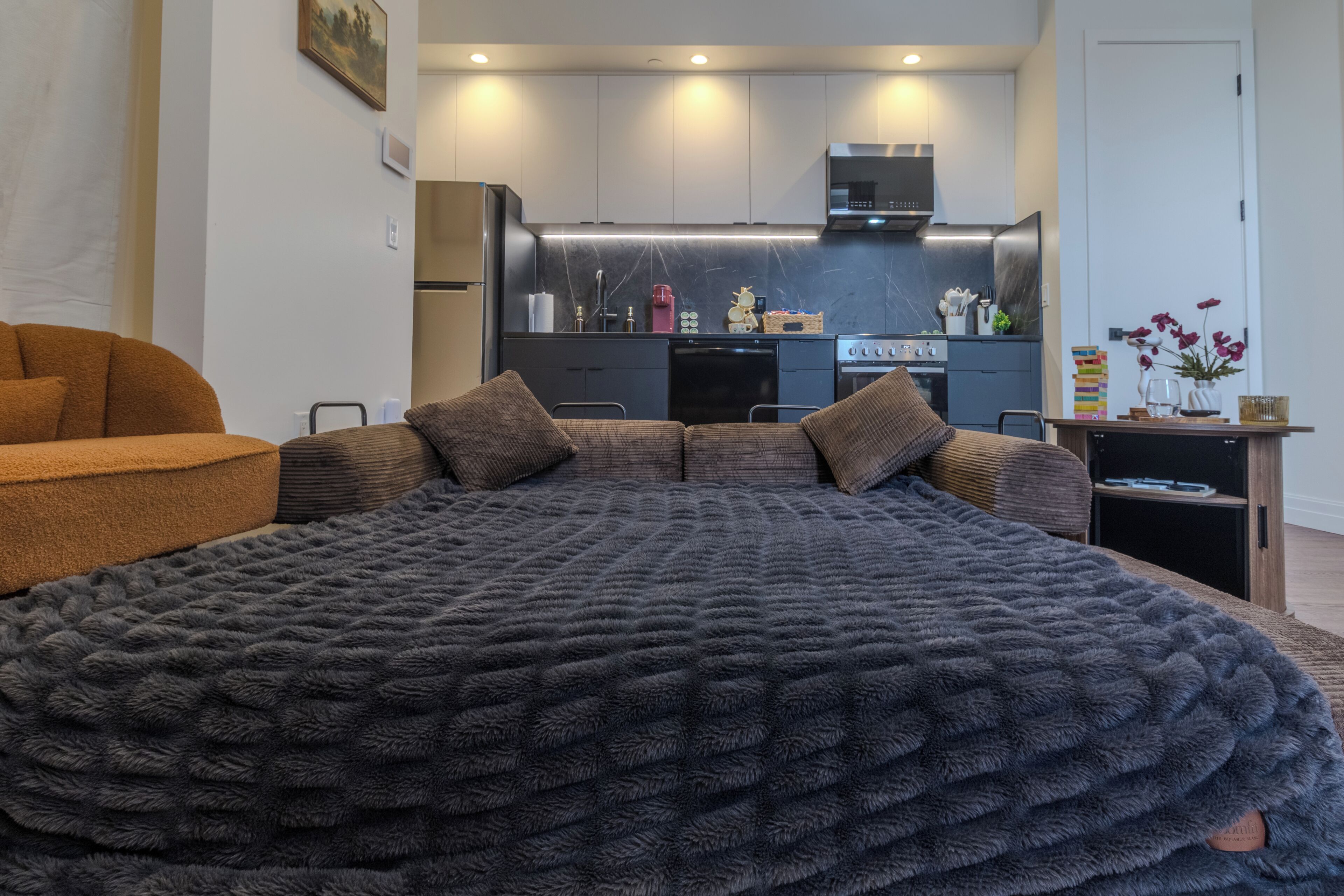 A bedroom with a large bed, a sofa, a kitchen in the background, and a door to the right.