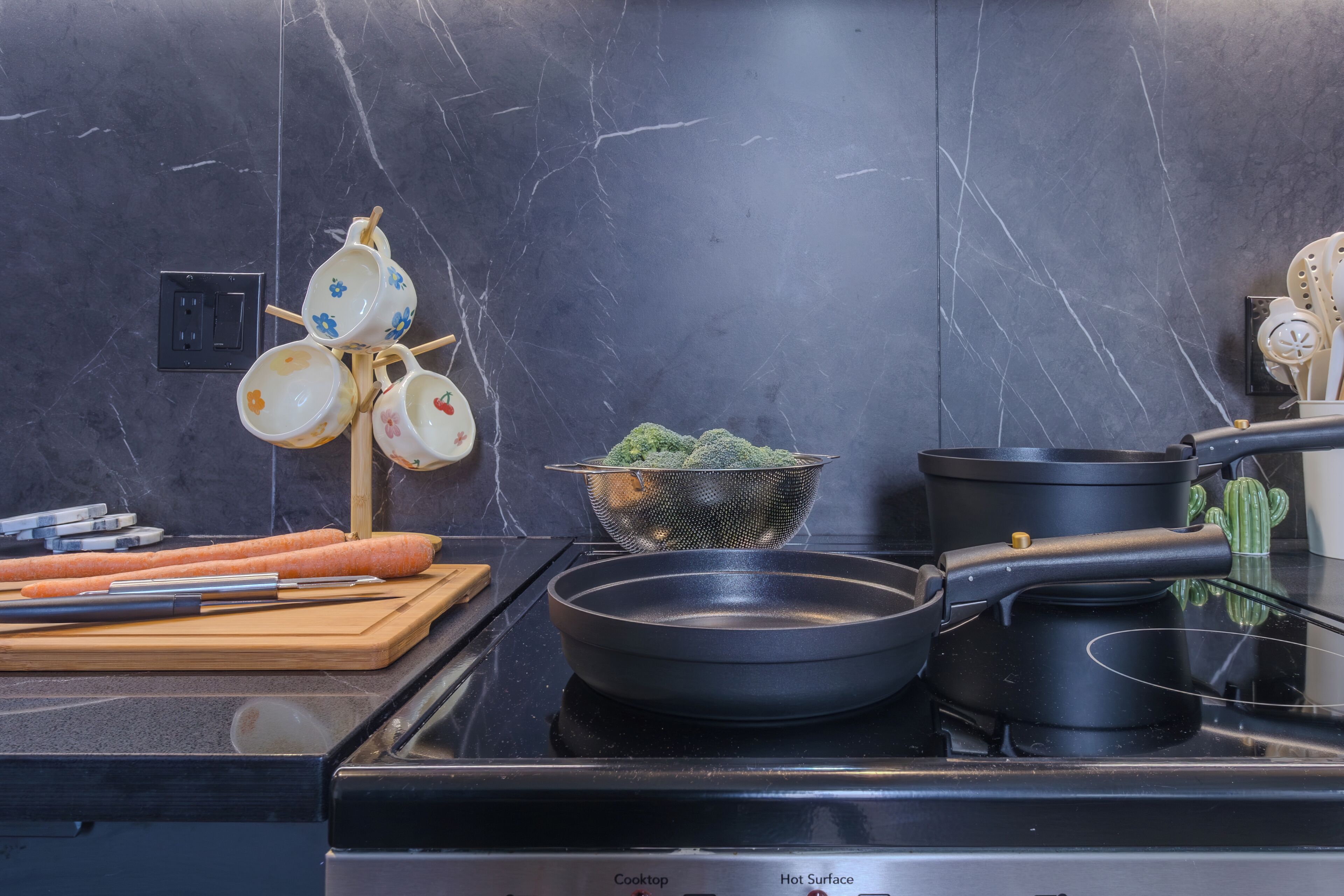 Une cuisine avec une plaque de cuisson noire, une planche à découper avec des carottes, une passoire et une casserole.