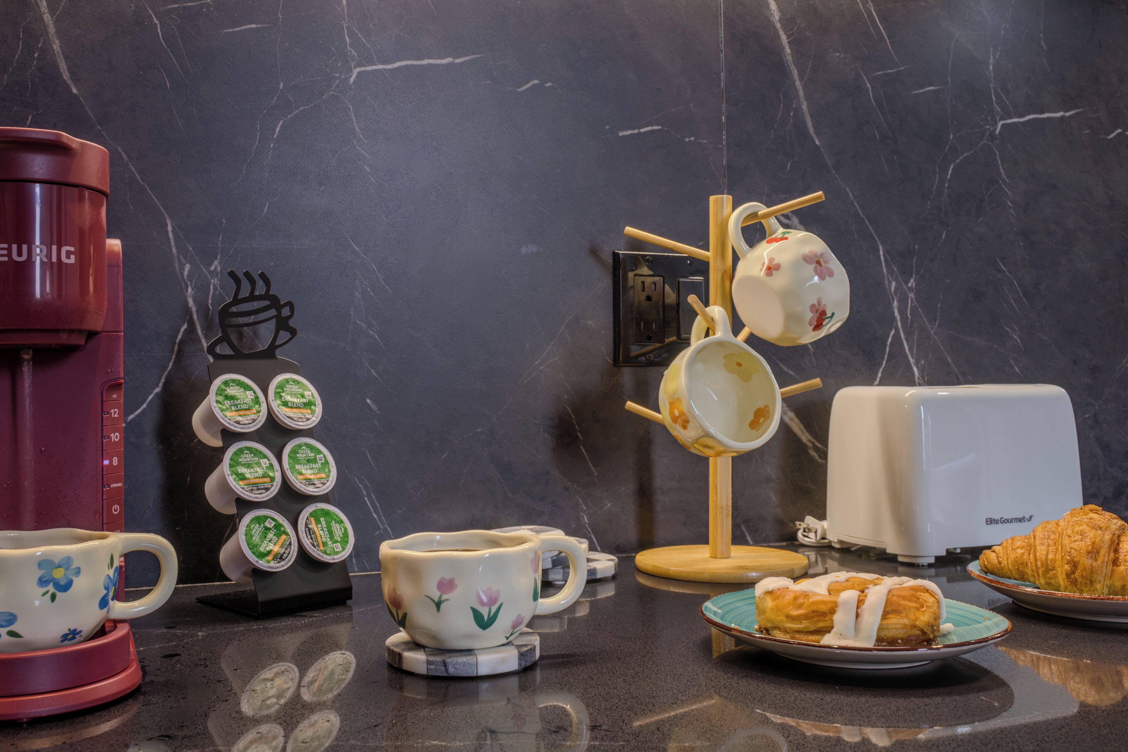 A kitchen counter with a coffee maker, teapot, cups, and a plate of pastries.