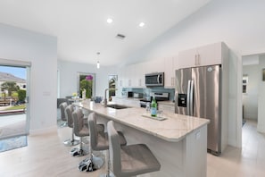 A modern kitchen with a central island, bar stools, stainless steel appliances, and a view of the outdoors. - Serene Waterfront Bliss – Private Pool Home Near Beach Access, Sleeps 8 (Marco Island)