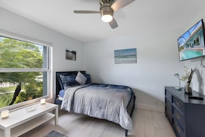A bedroom with a bed, a desk, a ceiling fan, and a TV on the wall.