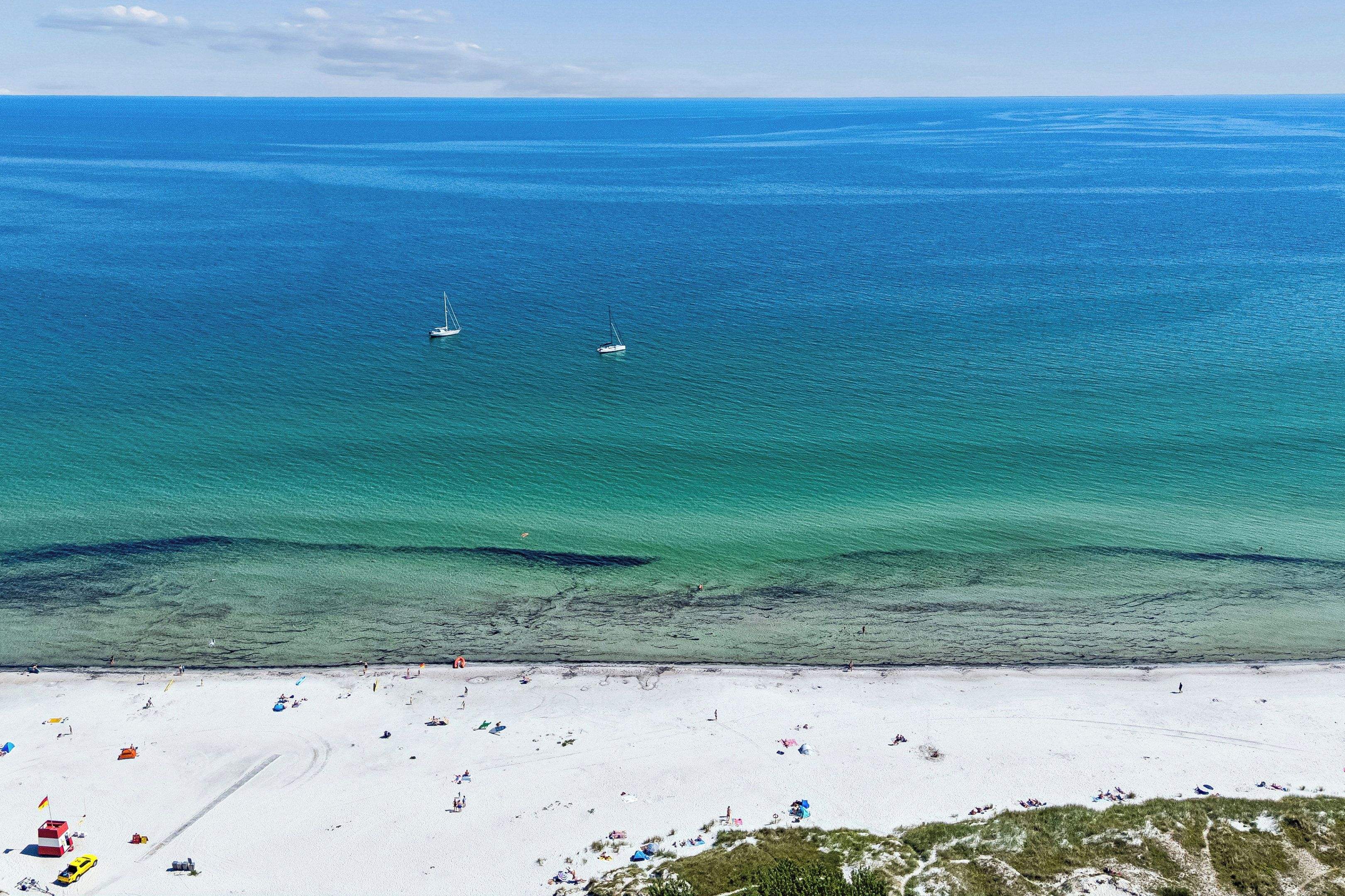 A beach with clear waters, two sailboats, and people sunbathing.