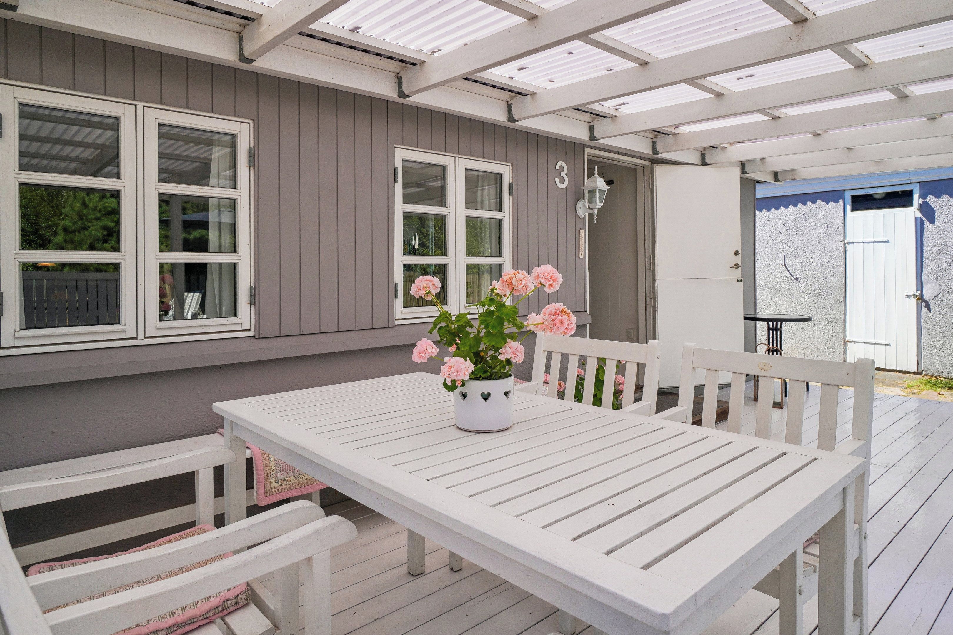 A covered patio with a white table and bench, a potted plant with flowers, and a house with grey siding and white trim.