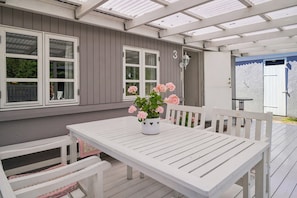 A covered patio with a white table and bench, a potted plant with flowers, and a house with grey siding and white trim.