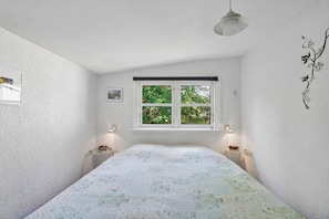 A bedroom with a bed, a window with a view of greenery, a wall-mounted light fixture, and a small shelf with a clock.