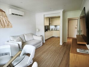 A modern living room with a sofa, chairs, a TV, and a kitchen area in the background. - Atlantic Ocean Flat | Stunning Ocean View in Cascais (Cascais)