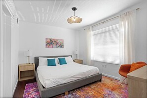 A bedroom with a bed, a desk, a chair, a painting, and a window with blinds. - 100 Steps to the Sand - Art Deco Miami Beach Apartment (Miami)