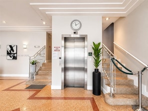 Un ascensor moderno con reloj digital, un letrero de advertencia y una planta en maceta junto a una escalera.