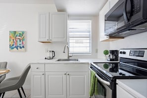 Una cocina compacta con muebles blancos, un lavamanos de acero inoxidable, una estufa negra y una ventana con persianas.