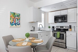 A modern kitchen with white cabinets, a stainless steel stove, a microwave, and a sink. There is a dining table with chairs and a small potted plant. - 100 Steps to the Sand - Art Deco Miami Beach Apartment (Miami)