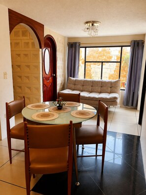 A dining area with a round glass table, four chairs, and a sofa in the background.