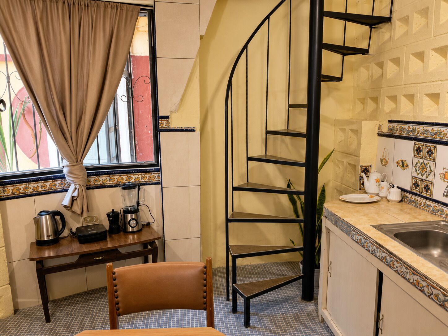 Una cocina con una escalera de caracol, una mesa de comedor con sillas, un ventanal con cortinas y un lavabo con azulejos decorativos.