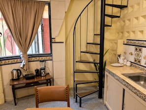 Una cocina con una escalera de caracol, una mesa de comedor con sillas, un ventanal con cortinas y un lavabo con azulejos decorativos.