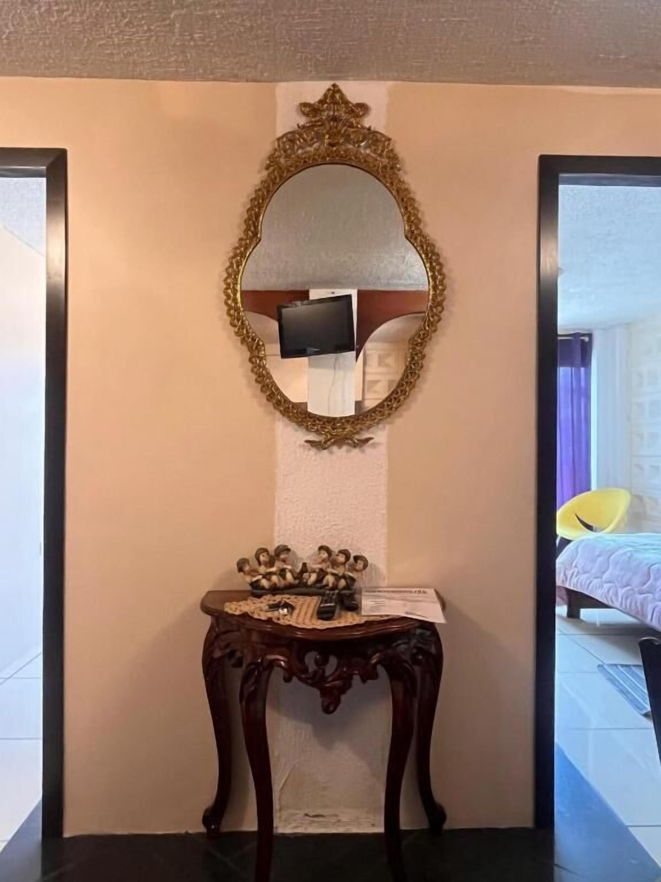 Habitación de hotel con un espejo grande y ornamentado, una pequeña consola de madera con objetos decorativos y una cama con colcha blanca.
