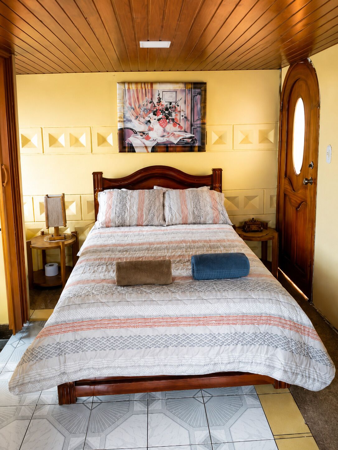 Une chambre avec un lit en bois, un tableau accroché au mur, une porte en bois et un sol à motifs.