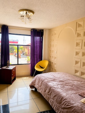 Un dormitorio con una cama, una silla, un aparador y una ventana con cortinas.