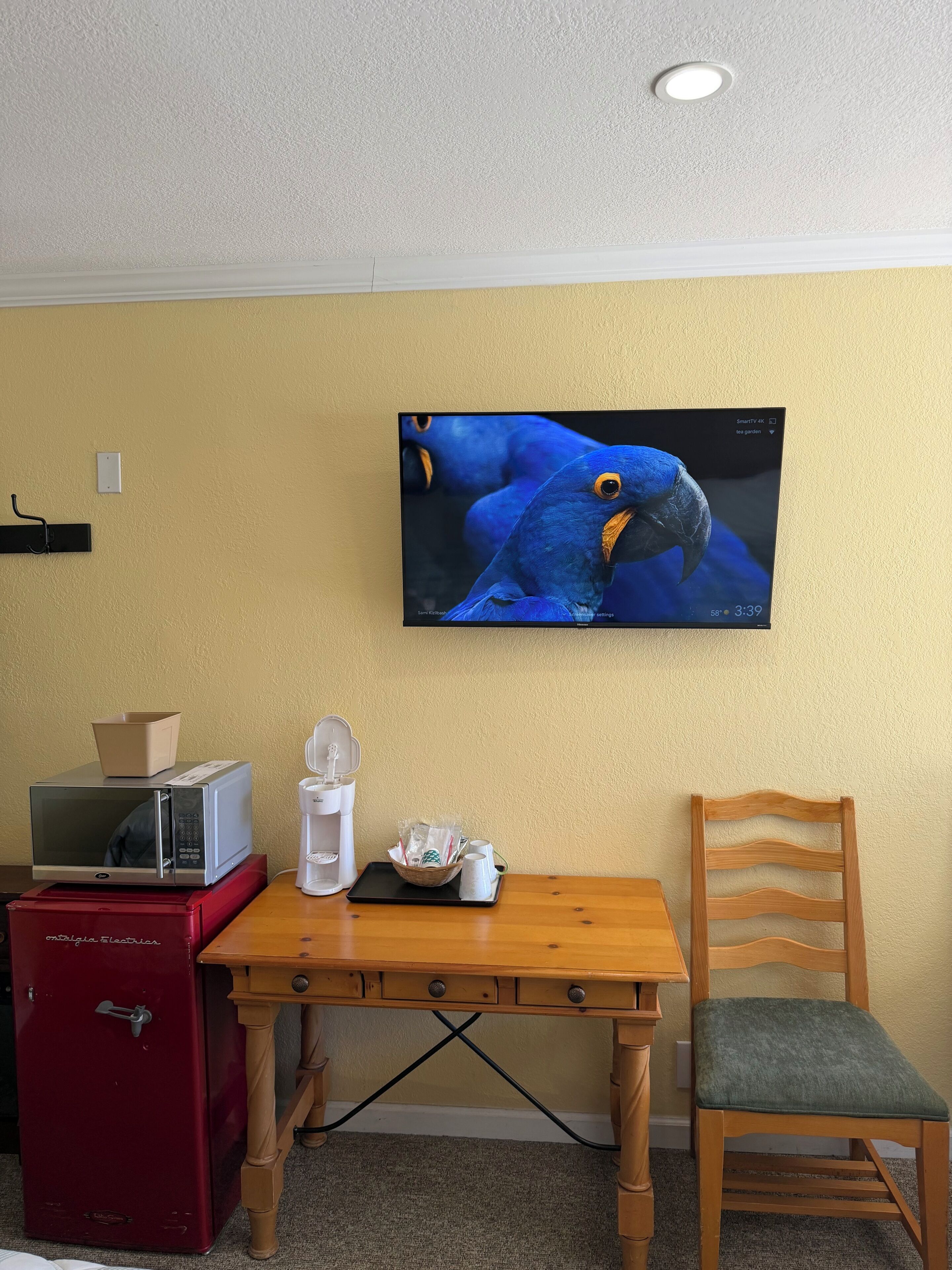 Habitación con un televisor montado en la pared mostrando un pájaro, un microondas en un mueble, una caja fuerte roja, una mesa de madera con una silla y una cesta con tazas.
