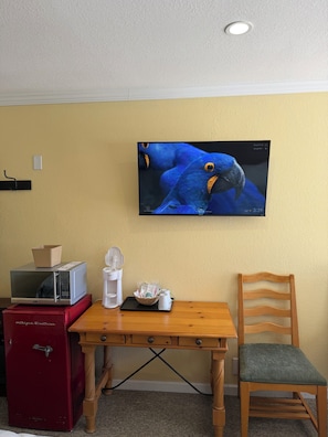 Habitación con un televisor montado en la pared mostrando un pájaro, un microondas en un mueble, una caja fuerte roja, una mesa de madera con una silla y una cesta con tazas.
